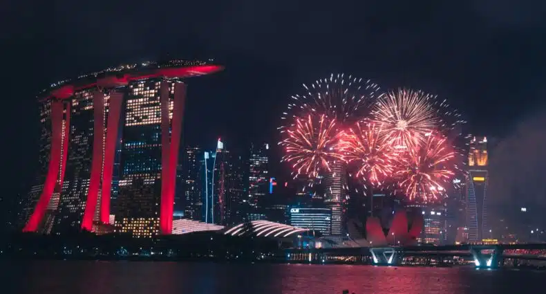 Fogos de artifício coloridos iluminam o céu noturno sobre Marina Bay Sands e prédios da cidade em Singapura, refletidos na água.