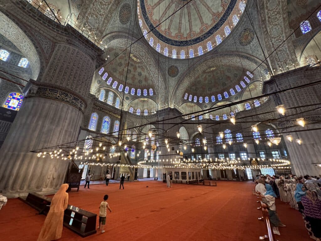 Interior de uma grande mesquita com tetos ornamentados, colunas, luzes penduradas e pessoas caminhando sobre um tapete vermelho. Essa é a Mesquita Azul, uma das opções de o que fazer em Istambul.
