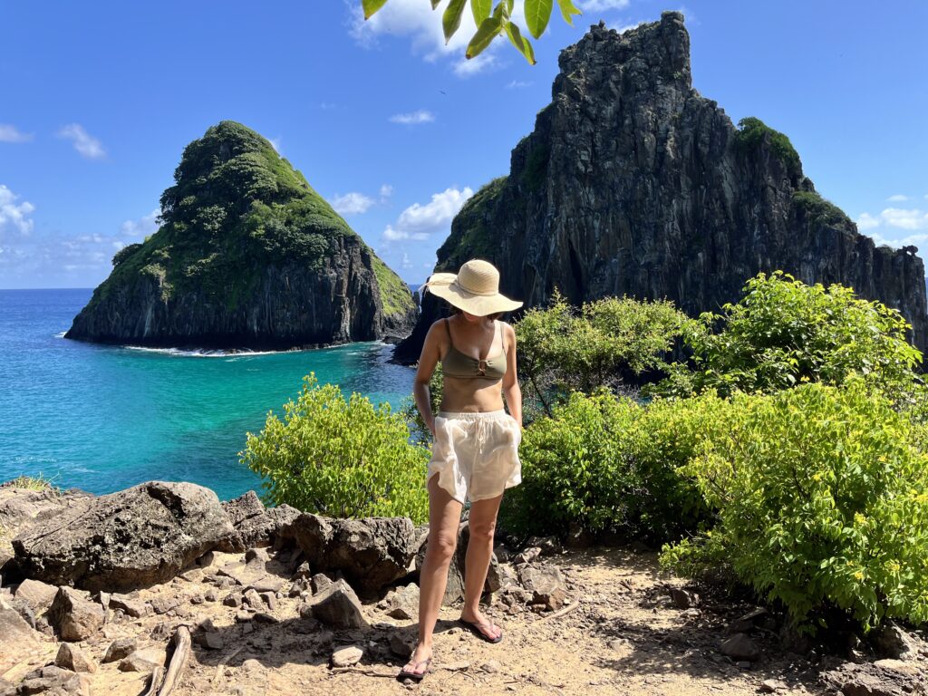 Uma mulher de chapéu está em um caminho rochoso perto de arbustos verdes, com grandes rochedos e águas azul-turquesa ao fundo.