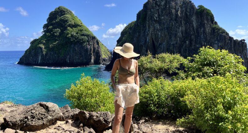 Uma mulher de chapéu está em um caminho rochoso perto de arbustos verdes, com grandes rochedos e águas azul-turquesa ao fundo.