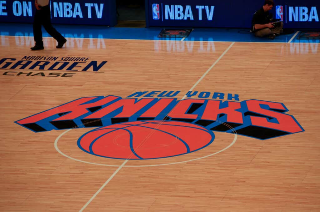 Logotipo do New York Knicks em uma quadra de basquete no Madison Square Garden com pessoas ao fundo.