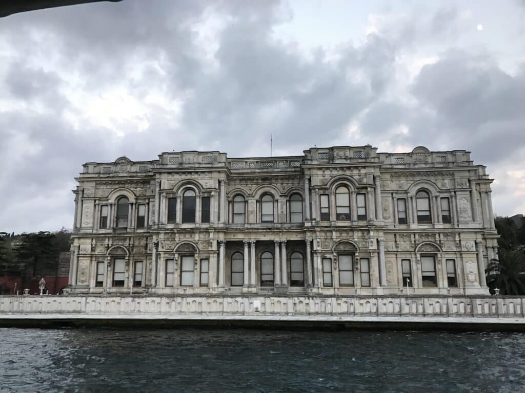 Um grande edifício histórico cinza, ornamentado, com janelas em arco, fica à beira-mar sob um céu nublado. Esse é o Palácio de Beylerbeyi, uma das opções de o que fazer em Istambul.