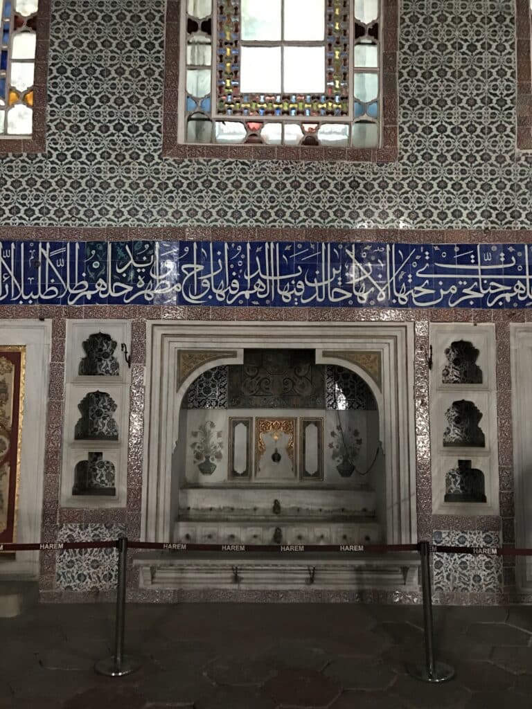 Sala ornamentada do Palácio Topkapi com azulejos de design intrincado, caligrafia árabe e uma janela acima de uma bacia de mármore e um nicho decorativo.