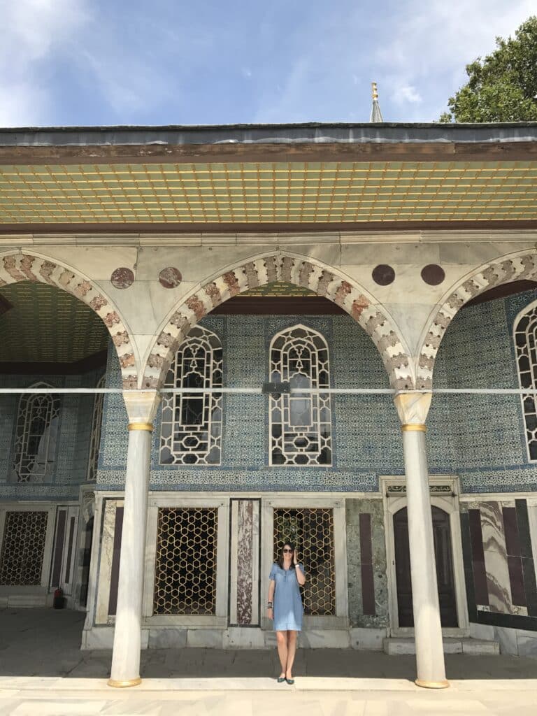 Uma mulher está sob arcos ornamentados em frente a um edifício histórico com azulejos intrincados e janelas de treliça. Esse é o Palácio Topkapi, uma das opções de o que fazer em Istambul.