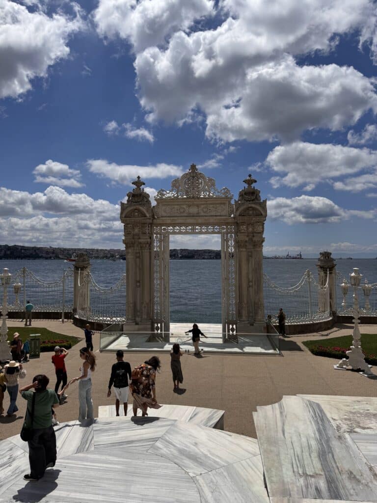 Portão ornamentado com vista para a água no Palácio Dolmabahçe, com pessoas de pé e caminhando em um dia ensolarado com nuvens dispersas.