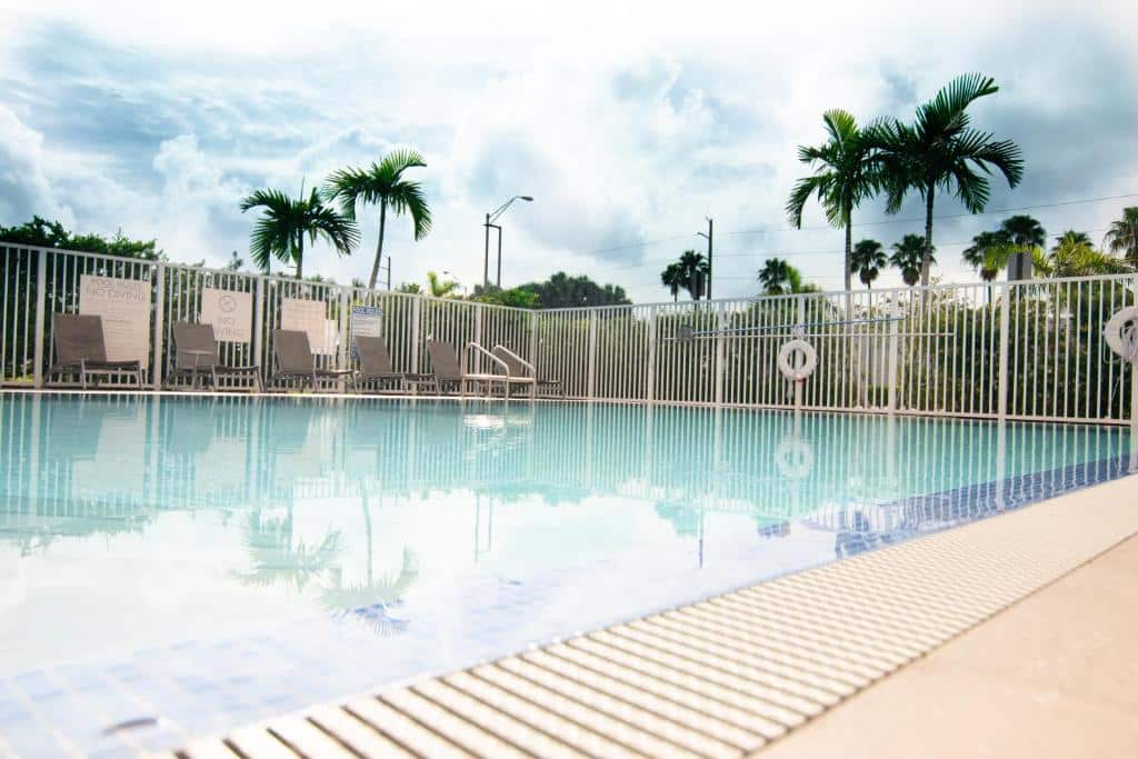 Piscina ao ar livre do Candlewood Suites Miami Intl Airport com espreguiçadeiras e palmeiras, cercada por uma cerca de metal branca sob um céu parcialmente nublado.