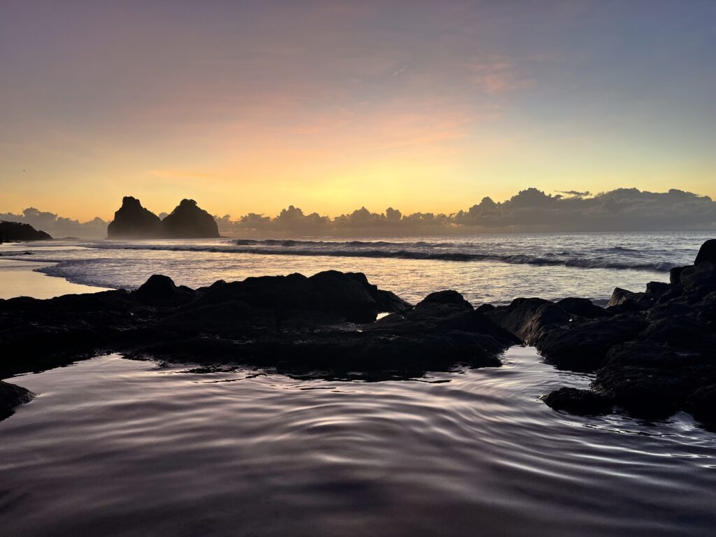 Litoral rochoso ao pôr do sol com águas calmas, silhueta de rochas e nuvens no horizonte sob um céu colorido.