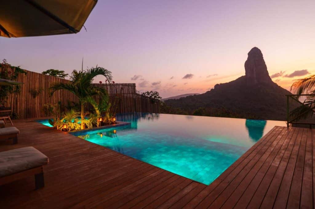 Uma piscina de borda infinita na Pousada Ecológica Akanã em um deck de madeira com vista para uma paisagem montanhosa ao pôr do sol, com a silhueta de um pico solitário ao fundo. Imagem para ilustrar post sobre onde ficar em Fernando de Noronha.