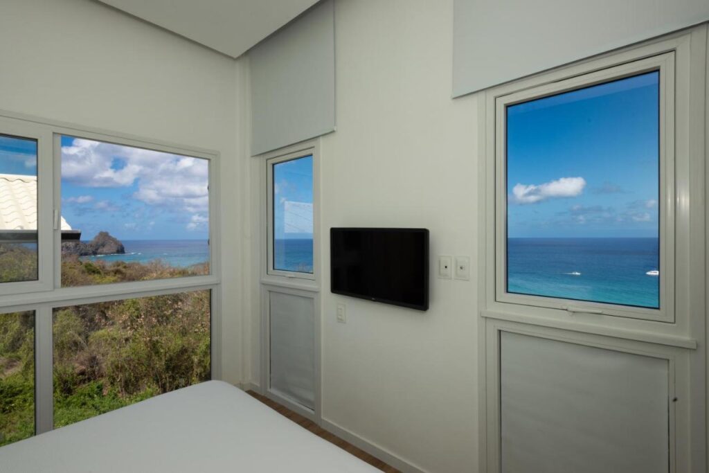 Quarto da Pousada Mar em Mim com janelas grandes mostrando uma vista do oceano e do céu. Uma TV está montada na parede entre as janelas.