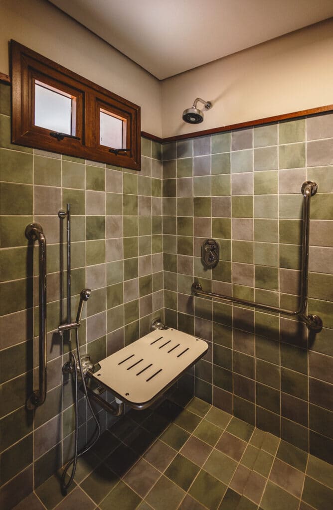 Chuveiro acessível da Pousada Moana com azulejos verdes, assento dobrável, barras de apoio e uma pequena janela na parte superior.