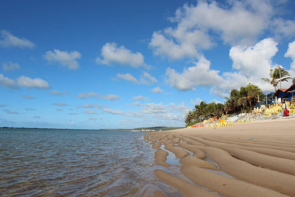 Praia Barra de São Miguel com a faixa de areia, cadeiras e residências no lado direito. No lado esquerdo está o mar com ondas suaves. A foto está no post de o que fazer na Barra de São Miguel.