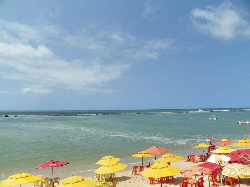 Praia Barra de São Miguel com guarda-sóis amarelos e vermelhos na areia, ondas azuis do oceano e um céu limpo.