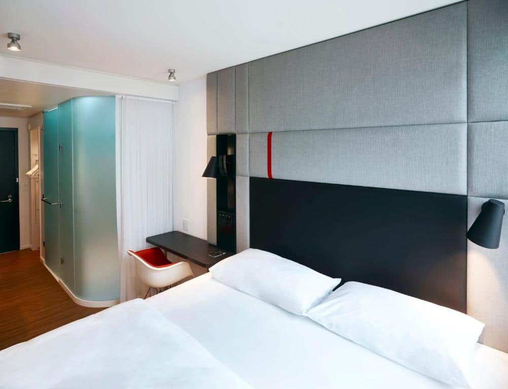 Quarto do citizenM Miami Brickell com uma cama grande, cabeceira cinza, detalhes em preto e vermelho, escrivaninha embutida e chuveiro de vidro fosco. Representa hotéis em Brickell.