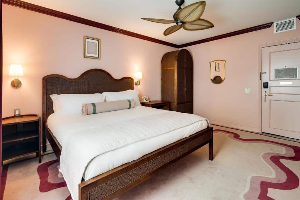 Quarto aconchegante do Esme Miami Beach com uma cama grande, almofadas decorativas, arandelas, um armário de madeira e ventilador de teto sobre um tapete rosa e branco. Representa Hotéis em South Beach, em Miami.