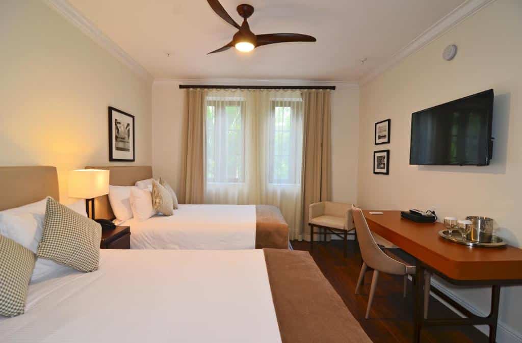 Quarto do Hotel St. Michel com duas camas de casal, ventilador de teto, TV montada na parede, escrivaninha com cadeira e janela grande com cortinas. Representa onde ficar em Miami.