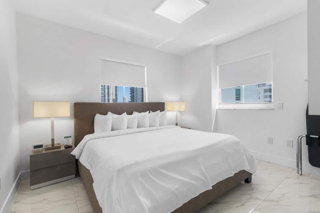 Um quarto minimalista do Fortune House Hotel Suites com uma cama grande, roupa de cama branca, duas mesas de cabeceira com luminárias e janelas com vista para a cidade. Representa hotéis em Brickell.