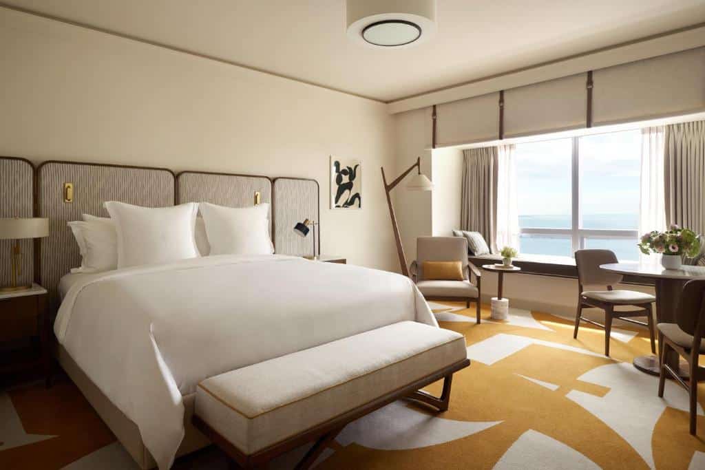 Um quarto do Four Seasons Hotel Miami com uma cama grande, banco, cadeiras e uma janela com vista para o mar. A decoração é moderna com tons neutros. Representa hotéis em Brickell.