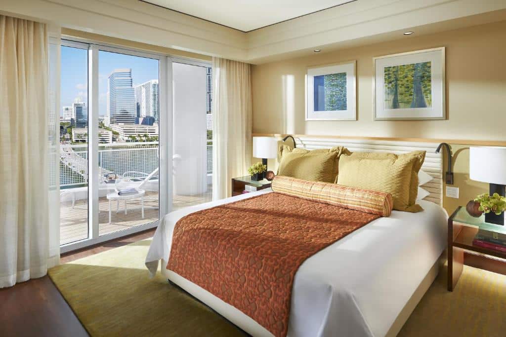 Quarto do Mandarin Oriental com uma cama grande, roupa de cama laranja e dois quadros emoldurados. Portas de vidro abrem para uma sacada com vista para a paisagem urbana. Representa onde ficar em Miami.