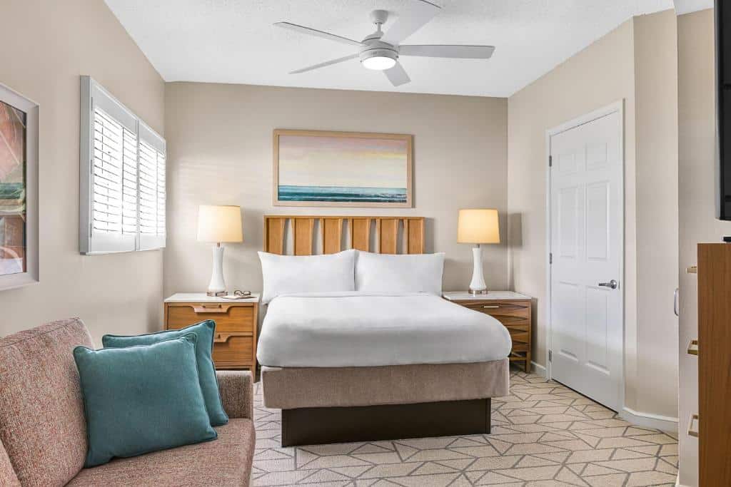 Um quarto aconchegante do Marriott’s Villas At Doral com uma cama de casal bem arrumada, duas luminárias, um ventilador de teto e arte na parede. Sofá com almofadas à esquerda. Representa onde ficar em Miami.