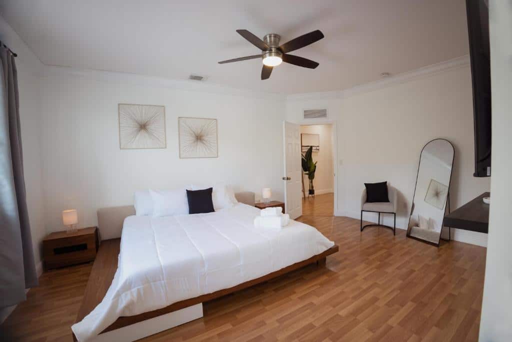 Quarto do Miami Luxury Villa Heated Pool & Pool Table com uma cama grande, ventilador de teto, piso de madeira, arte na parede, espelho, cadeira e uma porta que leva a outro cômodo. Representa onde ficar em Miami.