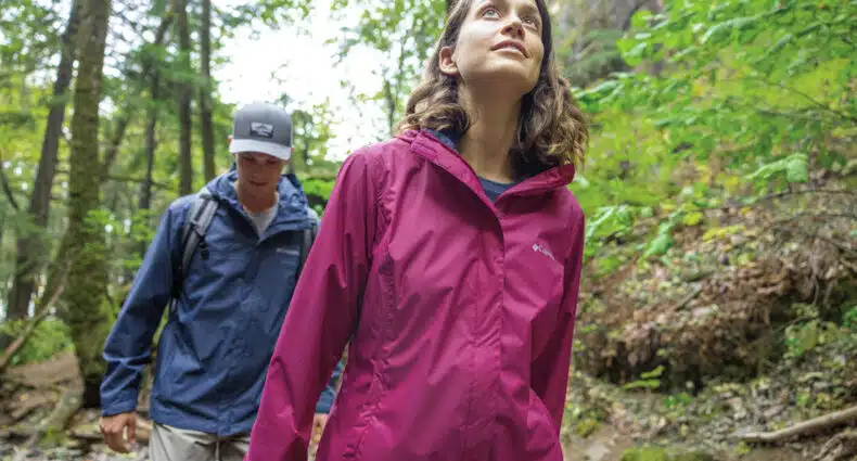Duas pessoas caminhando com roupa de trilha em uma floresta. A mulher na frente usa uma jaqueta vermelha, o homem atrás usa uma jaqueta azul e boné.