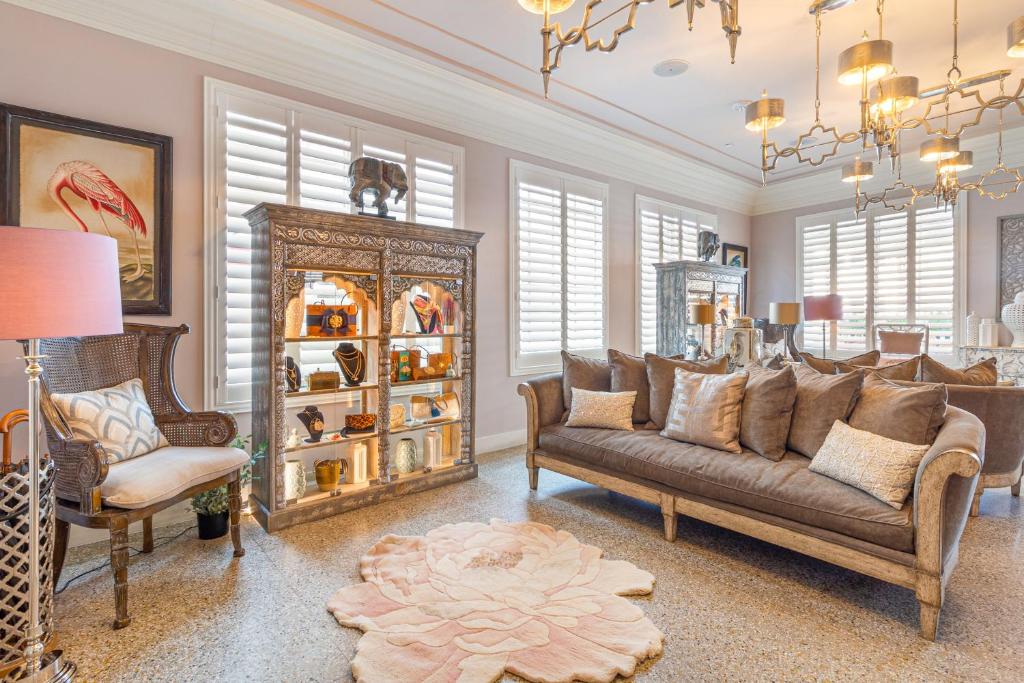 Sala de estar elegante do Henrosa Hotel com um sofá marrom, prateleiras decorativas, um tapete floral e vários lustres. Iluminada pelo sol através de janelas com venezianas.