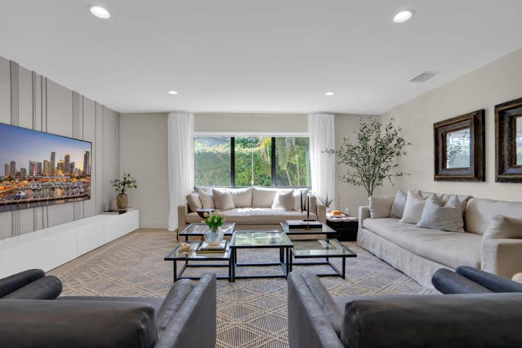 Sala de estar espaçosa do Miami GEM com dois sofás cinza, poltronas, mesas de centro, TV na parede, janela grande e plantas de interior.