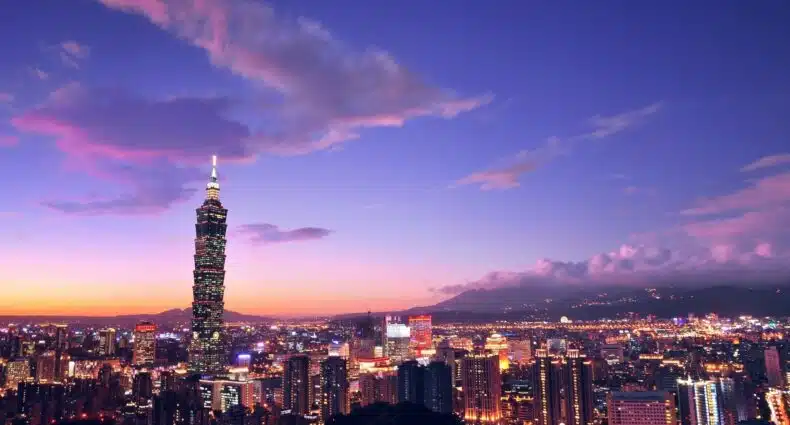 Um horizonte vibrante da cidade de Taipei ao anoitecer com um arranha-céu alto e iluminado (Taipei 101) e nuvens coloridas no céu.