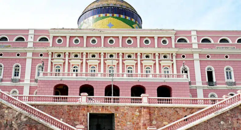 A imagem mostra um grande edifício do Teatro Amazonas, na cor rosa e branco com um telhado abobadado colorido, colunas decorativas e várias janelas.