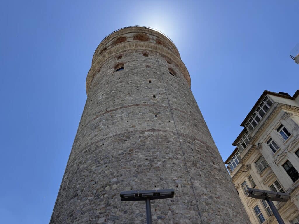 Uma torre alta e cilíndrica de pedra fica sob um céu azul claro, com a luz do sol brilhando ao redor do topo. Essa é a Torre Gálata, uma das opções de o que fazer em Istambul.