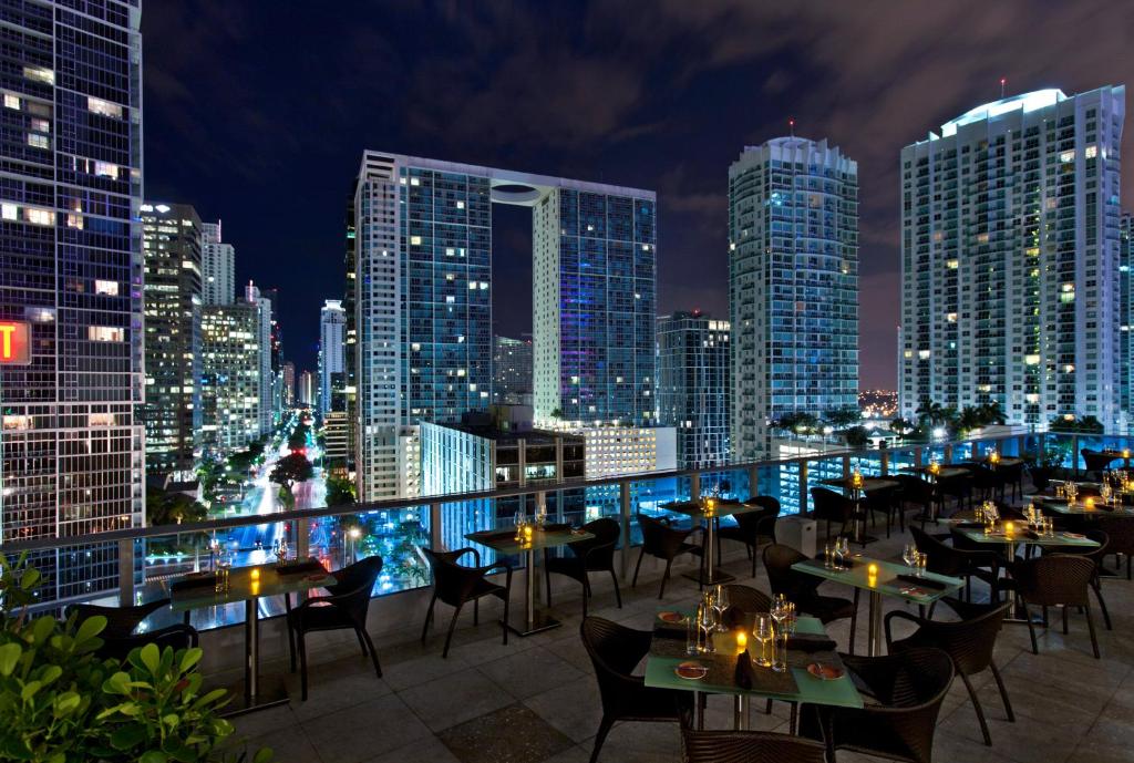 Vista da varanda do Kimpton EPIC Hotel, an IHG Hotel com horizonte da cidade à noite com edifícios altos e um restaurante na cobertura com mesas e cadeiras vazias. Representa onde ficar em Miami.