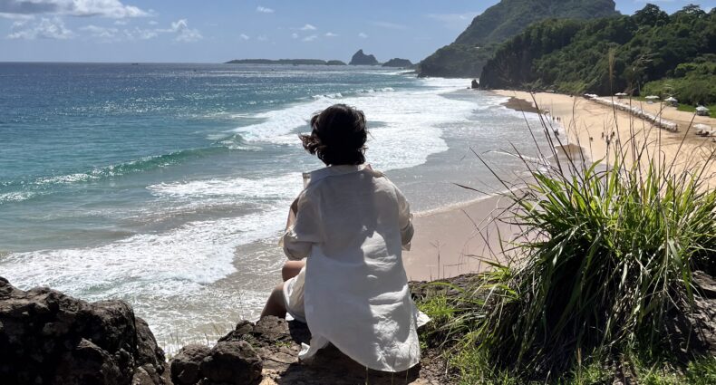 Uma pessoa de camisa branca está sentada em um penhasco rochoso com vista para uma praia arenosa, ondas do mar e uma montanha verde distante.