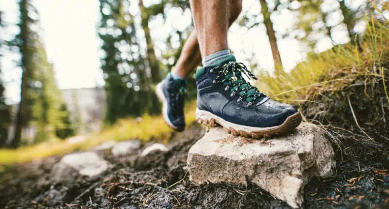 Uma pessoa usando botas Columbia de caminhada azuis pisa em uma pedra em uma trilha lamacenta na floresta, cercada por árvores e grama.