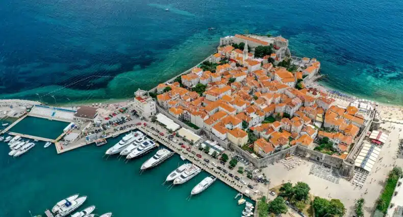 Vista aérea da cidade costeira, Budva, em Montenegro, com edifícios com telhados de terracota, uma marina com barcos e águas azuis cristalinas ao redor da área, para representar o que fazer em Montenegro.