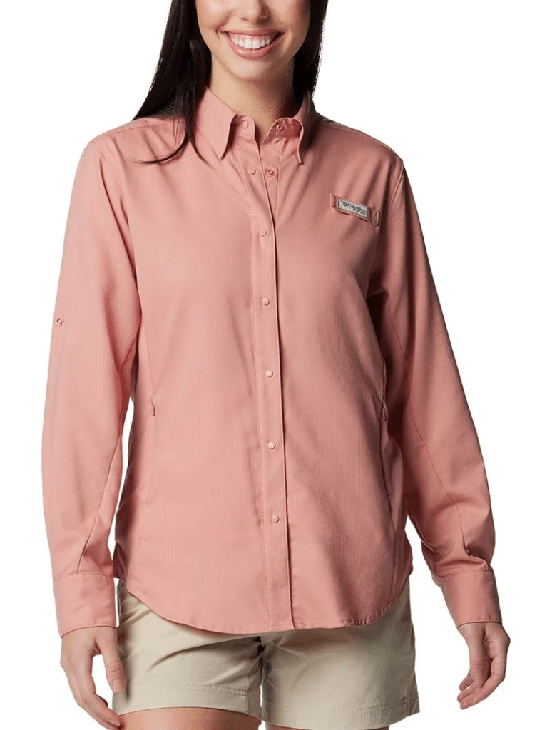 Pessoa vestindo uma camisa rosa de manga comprida com botões e shorts bege, sorrindo com as mãos apoiadas ao lado do corpo.