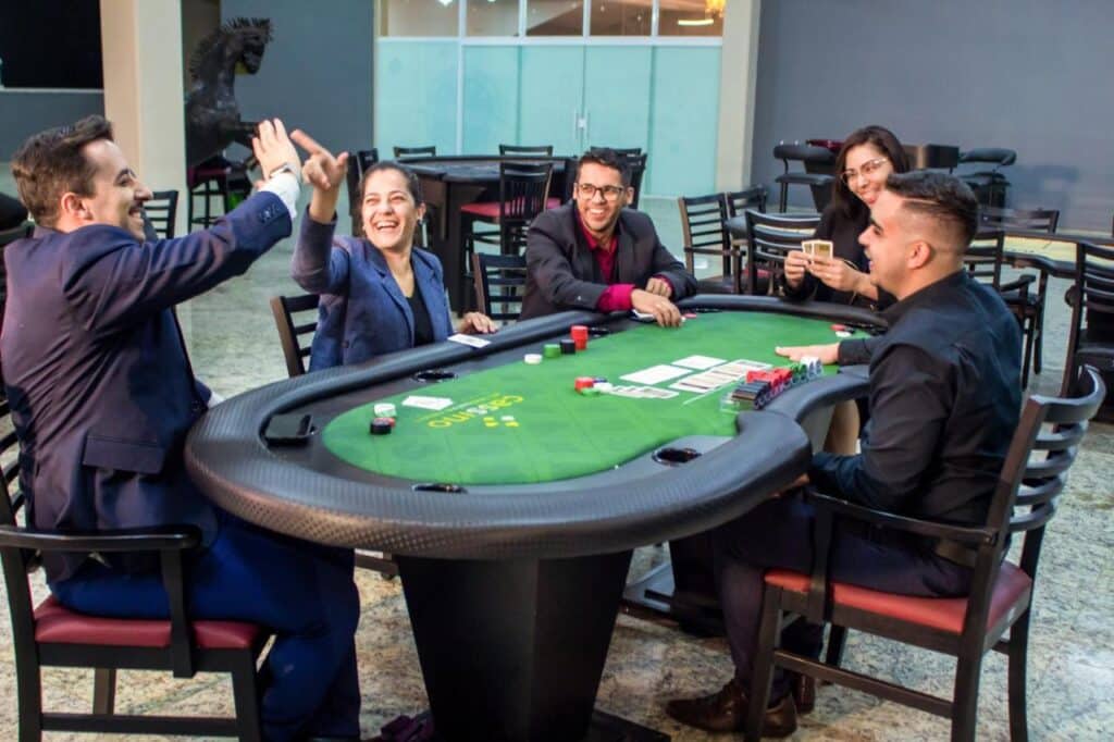 Cinco pessoas sentadas ao redor de uma mesa de pôquer em um ambiente animado no Cassino All Inclusive Resort Poços de Caldas, jogando cartas com fichas na mesa.