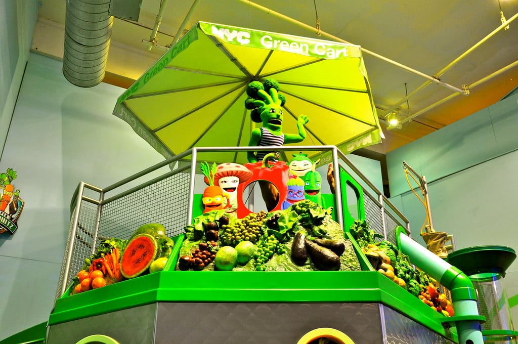 Exposição colorida com tema de frutas e vegetais, com cinco personagens de desenhos animados sob um guarda-chuva verde em uma plataforma elevada no Children's Museum de Manhattan.