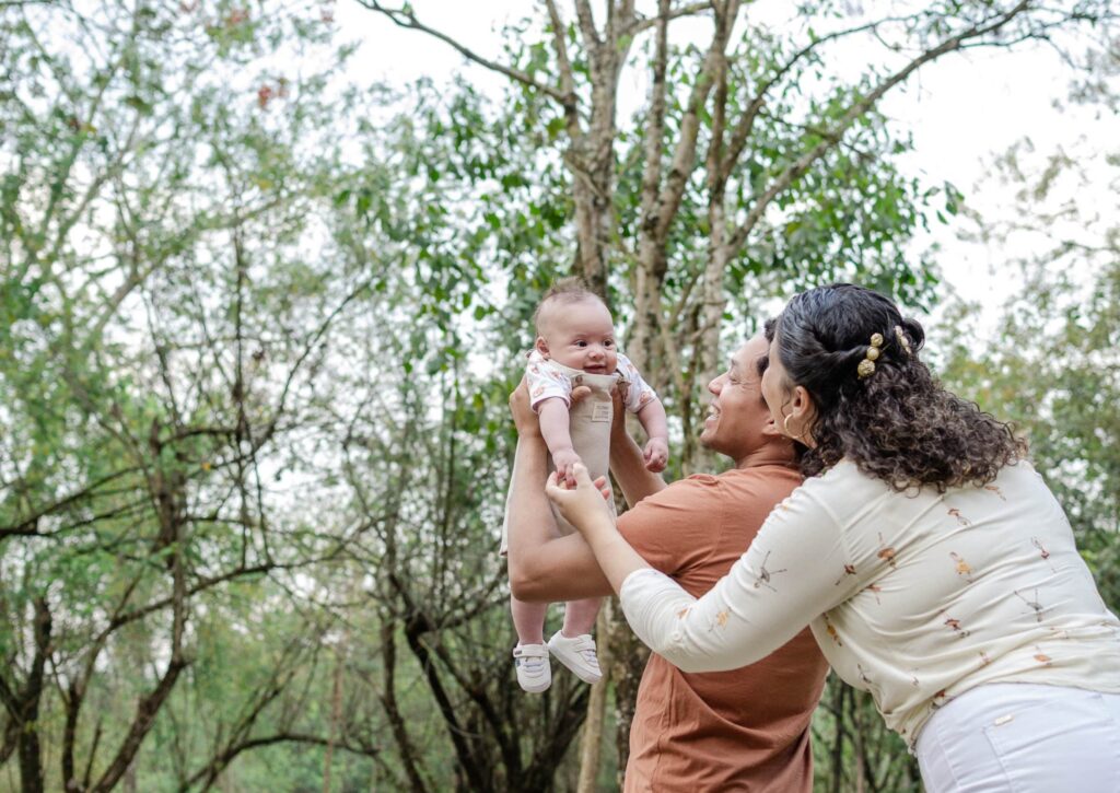 Um casal levanta um bebê sorridente no ar ao ar livre. Eles estão cercados por árvores e vegetação.