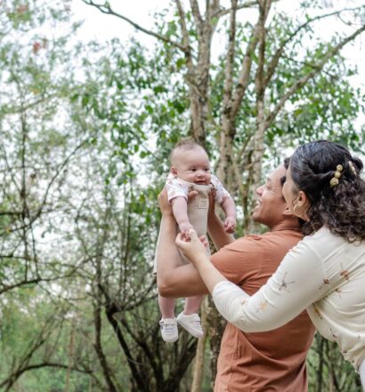 Um casal levanta um bebê sorridente no ar ao ar livre. Eles estão cercados por árvores e vegetação.