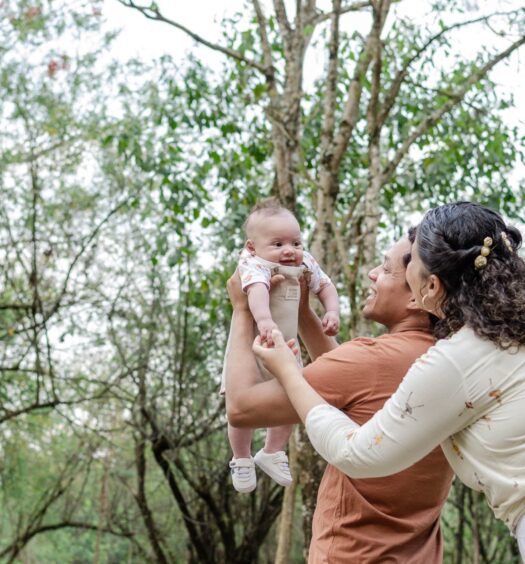 Um casal levanta um bebê sorridente no ar ao ar livre. Eles estão cercados por árvores e vegetação.