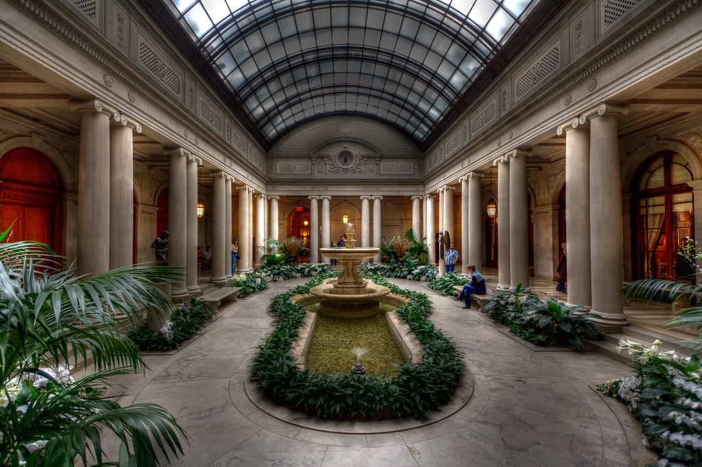 Pátio interno do Frick Collection com teto de vidro, fonte central, cercado por colunas e plantas verdes, com pessoas sentadas nas proximidades. Imagem para ilustrar post sobre museus em Nova York.
