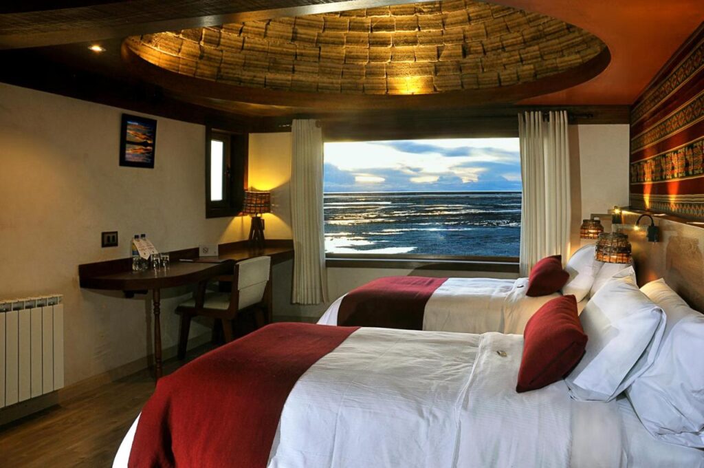 Quarto do Hotel Palacio de Sal com duas camas, detalhes em vermelho, uma escrivaninha e uma grande janela com vista para a praia e o oceano ao pôr do sol.