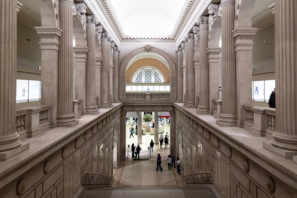 Interior do Metropolitan Museum of Art com colunas e arcos altos, pessoas caminhando no grande corredor abaixo e exposições iluminadas nas paredes. Imagem para ilustrar post sobre museus em Nova York.