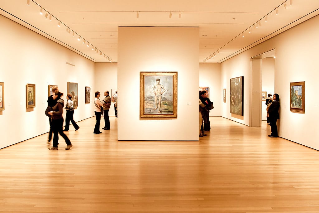 Pessoas observando pinturas no MoMa - Museum of Modern Art com paredes brancas e pisos de madeira. Imagem para ilustrar post sobre museus em Nova York.