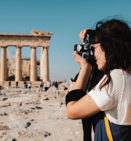 Uma mulher com uma mochila amarela tira uma foto de ruínas antigas sob um céu azul claro. Outra pessoa está de pé perto. Essa imagem ilustra o post sobre mochilão pela Europa.