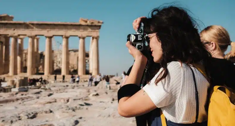 Uma mulher com uma mochila amarela tira uma foto de ruínas antigas sob um céu azul claro. Outra pessoa está de pé perto. Essa imagem ilustra o post sobre mochilão pela Europa.