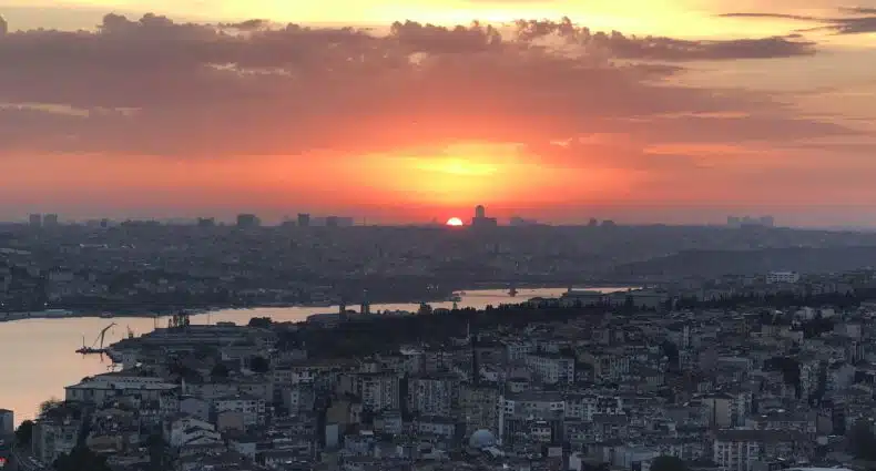 Uma paisagem urbana ao pôr do sol, com o sol se pondo no horizonte, edifícios e água visíveis em primeiro plano e nuvens acima. A imagem ilustra o posto sobre o que fazer em Istambul.