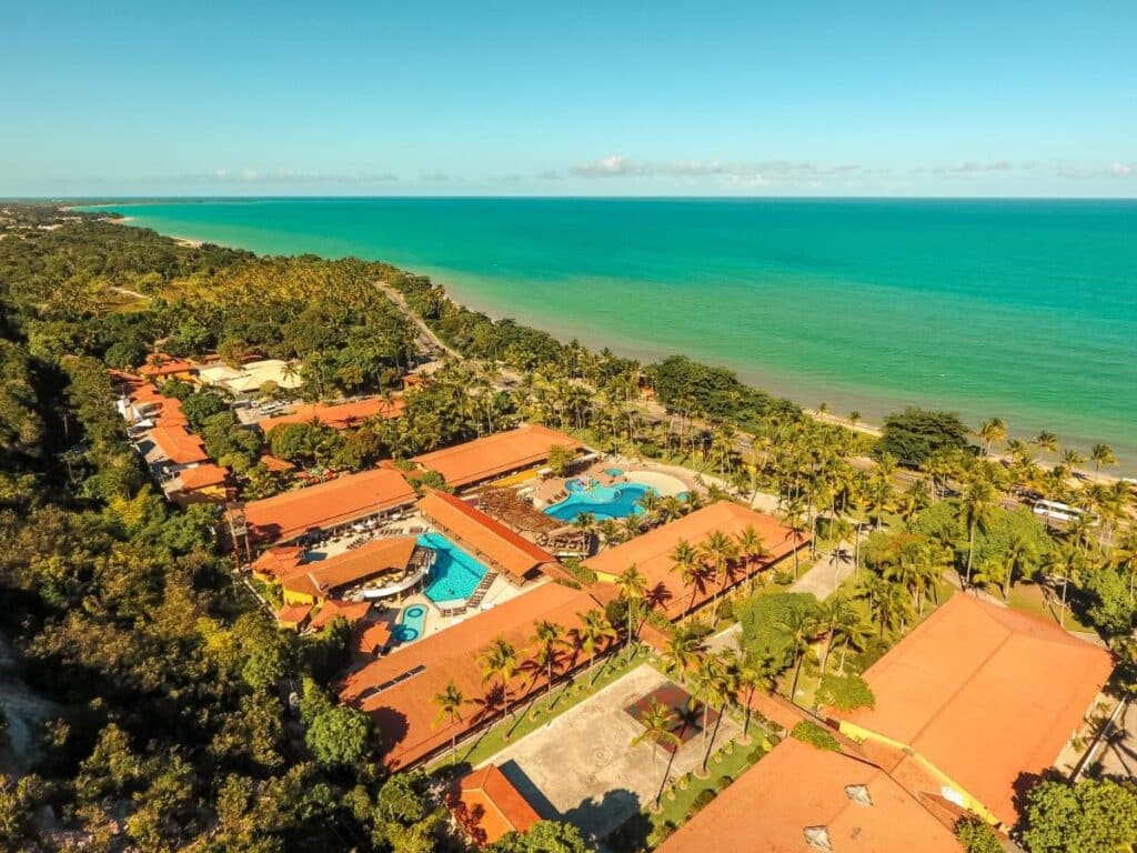 Vista aérea do Porto Seguro Praia Resort - All Inclusive com telhados vermelhos, piscinas e palmeiras ao longo de uma longa praia de areia e oceano azul-turquesa.