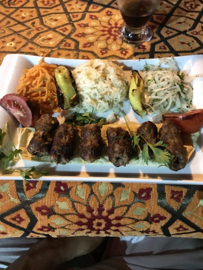 Um prato de espetinhos de carne grelhada com acompanhamentos de vegetais grelhados, repolho picado, cebola fatiada e uma fatia de tomate sobre uma toalha de mesa estampada.