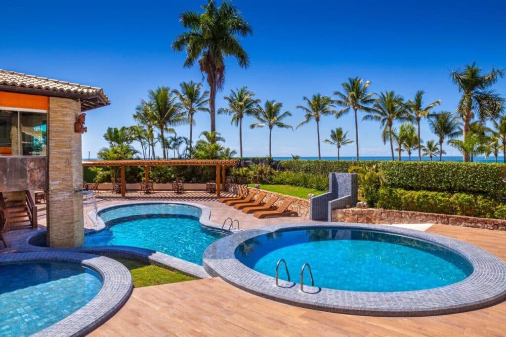 Área de piscina ao ar livre da Quinta do Sol Praia Hotel com duas piscinas redondas, cercadas por palmeiras e espreguiçadeiras, sob um céu azul claro.